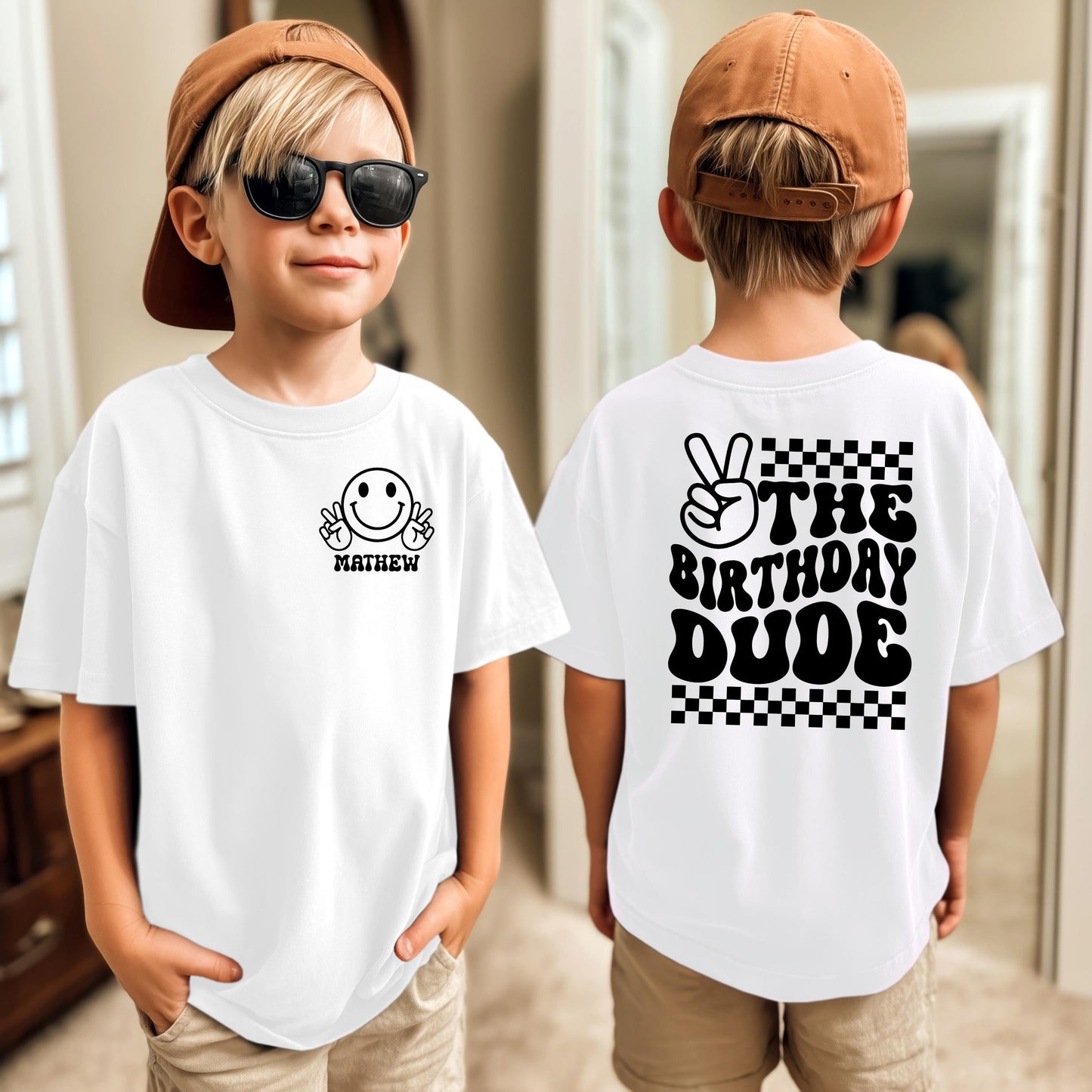 Custom birthday t-shirt design featuring a playful smiley face with peace signs on the front chest area, with space below to personalize with a name or custom text. The back of the shirt has a bold retro-style design that reads “THE BIRTHDAY DUDE” in chunky lettering, paired with a peace sign hand graphic and a checkerboard pattern for a fun, celebratory vibe. Perfect for kids’ or adults’ birthday outfits with custom personalization options.