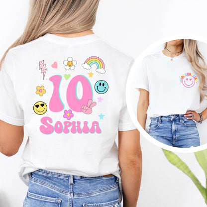 birthday T-shirt with colorful designs. The front of the shirt features a pink smiley face with the words “BIRTHDAY GIRL” above it in rainbow colors. The back of the shirt displays age in a large number surrounded by cute doodles like flowers, hearts, stars, a rainbow, and smiley faces, along with a customizable name (e.g., “Emma”). The age and name on the shirt can be personalized.