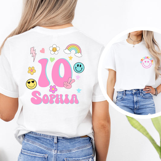 birthday T-shirt with colorful designs. The front of the shirt features a pink smiley face with the words “BIRTHDAY GIRL” above it in rainbow colors. The back of the shirt displays age in a large number surrounded by cute doodles like flowers, hearts, stars, a rainbow, and smiley faces, along with a customizable name (e.g., “Emma”). The age and name on the shirt can be personalized.