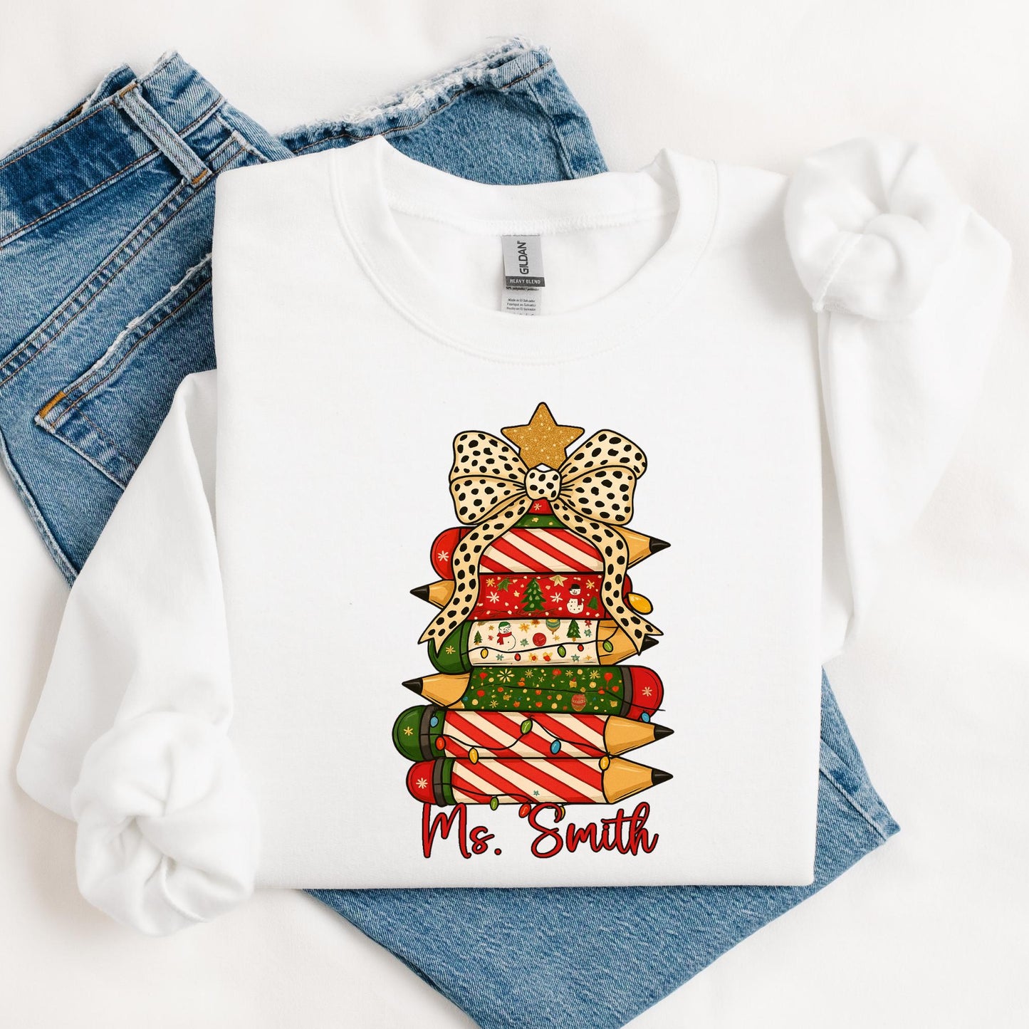 white sweatshirt featuring a festive Christmas design of stacked colored pencils decorated in red, green, and holiday patterns arranged like a Christmas tree. The tree is topped with a gold star and a polka-dot bow, with the text “Ms. Smith” written in red script below the design. The woman is seated casually, smiling, with light-wash ripped jeans and cozy socks.