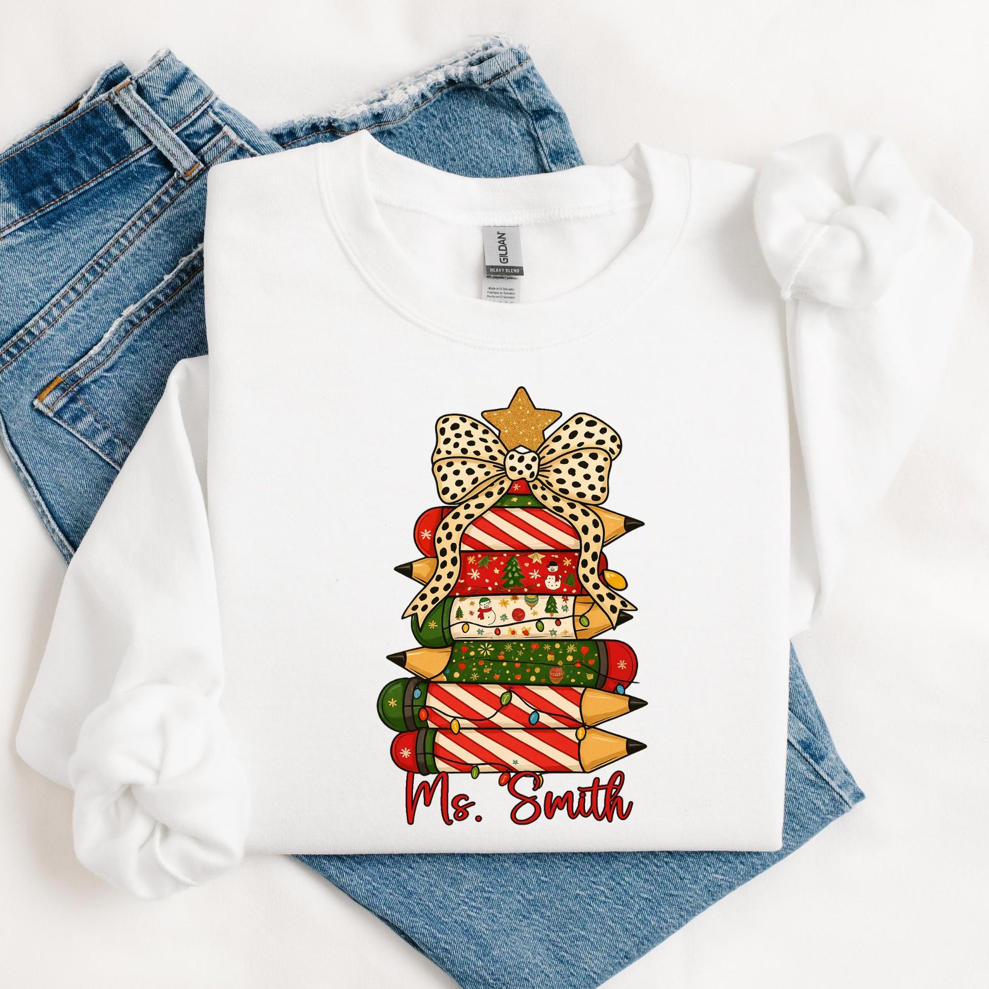 white sweatshirt featuring a festive Christmas design of stacked colored pencils decorated in red, green, and holiday patterns arranged like a Christmas tree. The tree is topped with a gold star and a polka-dot bow, with the text “Ms. Smith” written in red script below the design. The woman is seated casually, smiling, with light-wash ripped jeans and cozy socks.