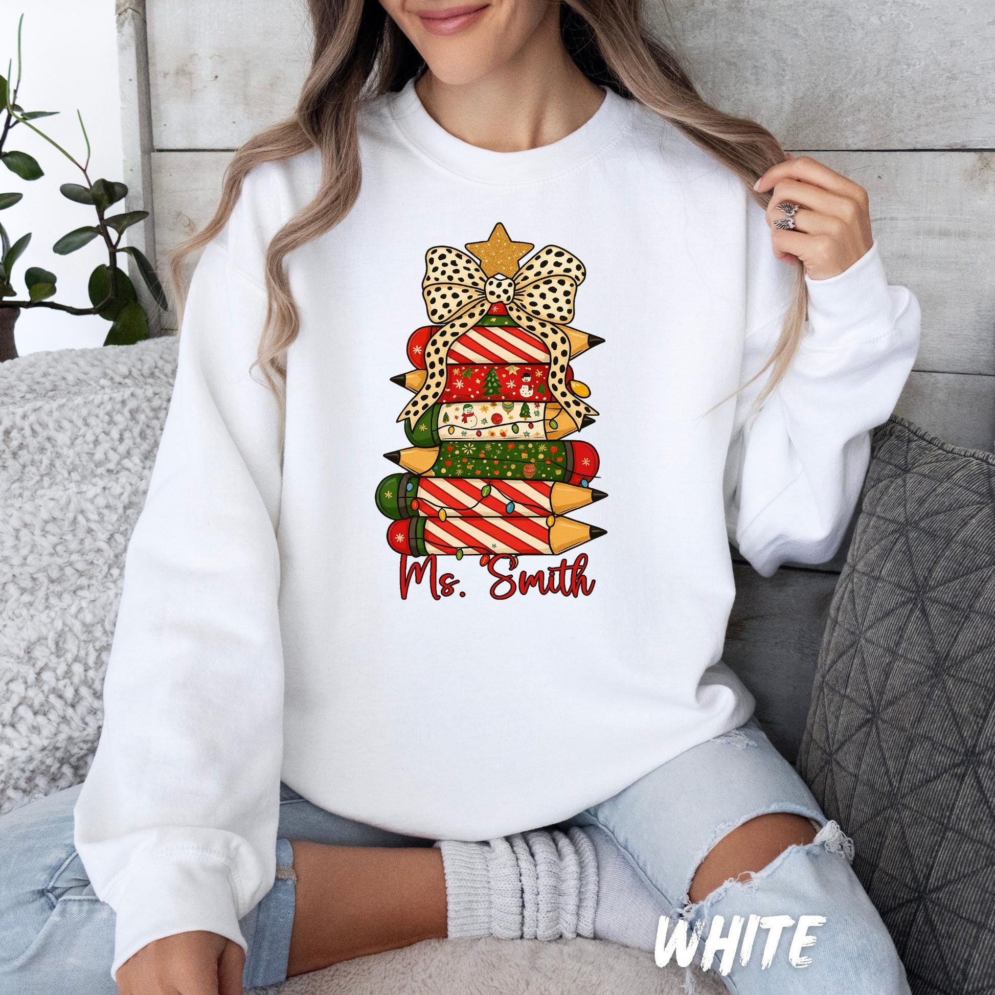 sweatshirt featuring a festive Christmas design of stacked colored pencils decorated in red, green, and holiday patterns arranged like a Christmas tree. The tree is topped with a gold star and a polka-dot bow, with the text “Ms. Smith” written in red script below the design. The woman is seated casually, smiling, with light-wash ripped jeans and cozy socks.