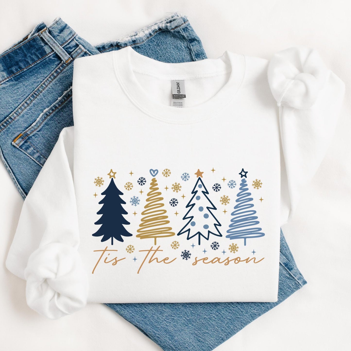 White Christmas sweatshirt featuring four minimalist Christmas trees in navy blue, gold, and light blue with snowflakes and stars. The text “‘Tis the Season” is written in elegant script. Cozy unisex holiday crewneck, perfect for winter outfits, Christmas gifts, or festive family photos.