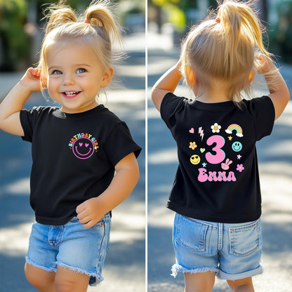 A smiling toddler wearing a black birthday T-shirt with colorful designs. The front of the shirt features a pink smiley face with the words “BIRTHDAY GIRL” above it in rainbow colors. The back of the shirt displays a large number “3,” surrounded by cute doodles like flowers, hearts, stars, a rainbow, and smiley faces, along with a customizable name (e.g., “Emma”). The age and name on the shirt can be personalized.