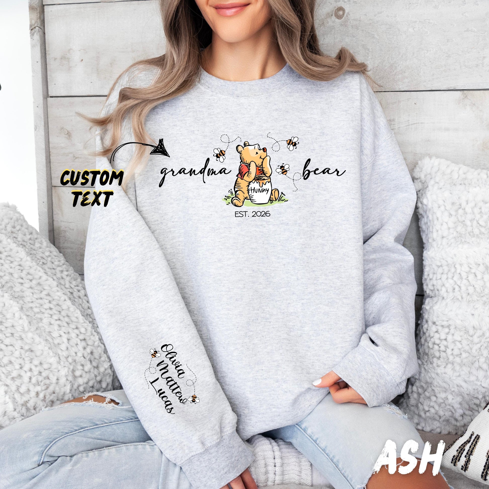 A woman wears a sweatshirt featuring a cute illustration of a bear holding a "hunny" pot surrounded by bees and flowers. The text around the bear reads "grandma bear" with "EST. 2026" below. The left sleeve displays customizable names of children - with small bees flying between them. The design allows personalization of the family member title (e.g., mama, nonna, auntie) and children's names, as well as the "Est." year.