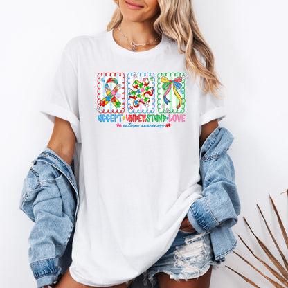 Autism awareness shirt with colorful puzzle ribbon, puzzle piece, and bow design, featuring the phrase “Accept Understand Love” on a white t-shirt, worn casually with denim jacket and shorts.