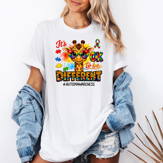 Autism awareness t-shirt featuring a cute giraffe wearing colorful heart-shaped glasses with puzzle pieces and the phrase “It’s OK to be Different #AutismAwareness” printed on a white shirt worn with denim jacket and shorts.