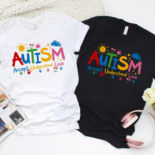 Autism awareness shirt with colorful “AUTISM Accept Understand Love” design featuring sun, flowers, stars, and handprints on white and black t-shirts flat lay.