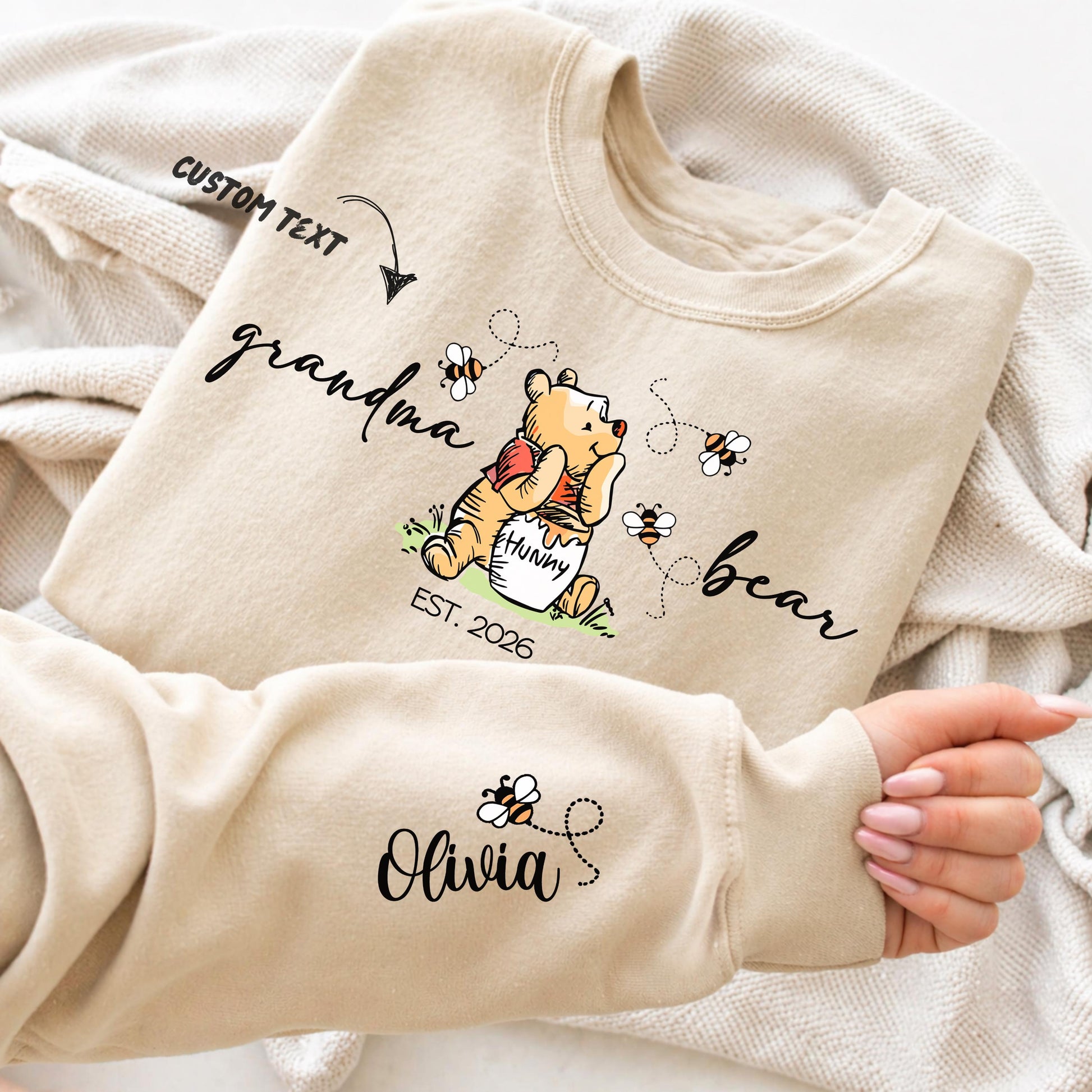 A woman wears a sweatshirt featuring a cute illustration of a bear holding a "hunny" pot surrounded by bees and flowers. The text around the bear reads "grandma bear" with "EST. 2026" below. The left sleeve displays customizable names of children - with small bees flying between them. The design allows personalization of the family member title (e.g., mama, nonna, auntie) and children's names, as well as the "Est." year.