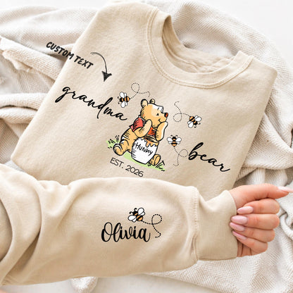 A woman wears a sweatshirt featuring a cute illustration of a bear holding a "hunny" pot surrounded by bees and flowers. The text around the bear reads "grandma bear" with "EST. 2026" below. The left sleeve displays customizable names of children - with small bees flying between them. The design allows personalization of the family member title (e.g., mama, nonna, auntie) and children's names, as well as the "Est." year.