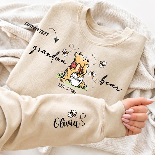 A woman wears a sweatshirt featuring a cute illustration of a bear holding a "hunny" pot surrounded by bees and flowers. The text around the bear reads "grandma bear" with "EST. 2026" below. The left sleeve displays customizable names of children - with small bees flying between them. The design allows personalization of the family member title (e.g., mama, nonna, auntie) and children's names, as well as the "Est." year.