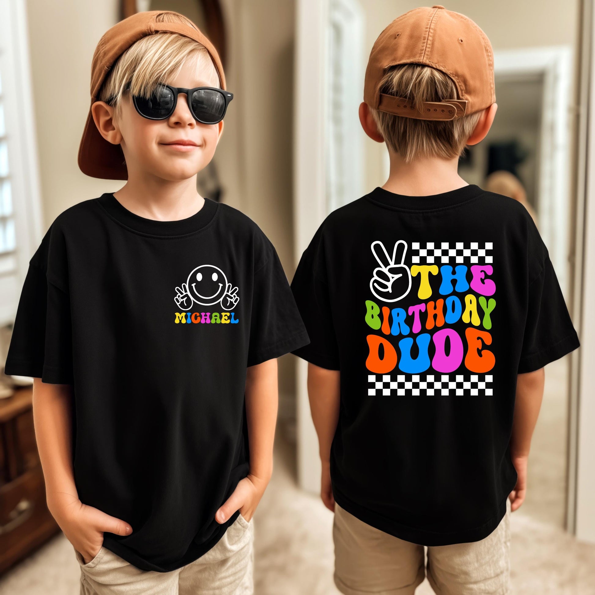 May include: Black t-shirt with a white smiley face and the name "Sebastian" printed on the front. The back of the shirt features the text "The Birthday Dude" with a peace sign and checkered borders. The child is wearing a brown baseball cap and sunglasses.