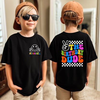 May include: Black t-shirt with a white smiley face and the name "Sebastian" printed on the front. The back of the shirt features the text "The Birthday Dude" with a peace sign and checkered borders. The child is wearing a brown baseball cap and sunglasses.