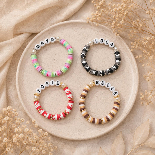 Personalized friendship bracelets displayed on a neutral ceramic plate with a soft beige aesthetic background. Four custom name bracelets feature colorful clay beads in Candy, Cookies & Cream, Peppermint Twist, and Caramel Latte color combinations, accented with pearl beads and silver or gold spacers. Handmade elastic bracelets make thoughtful gifts for best friends, sisters, daughters, birthdays, and special occasions.