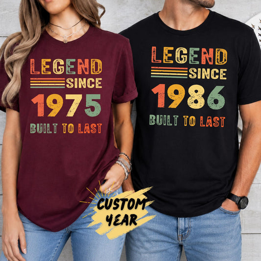A man wearing a black unisex t-shirt and a woman wearing a maroon unisex softstyle t-shirt featuring a customizable vintage "Legend Since" birthday design with retro colors and personalized birth year.
