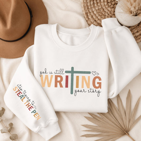White Gildan crewneck sweatshirt featuring the inspirational "God Is Still Writing Your Story" design with a cross and colorful typography, plus a matching faith message printed on the sleeve.
