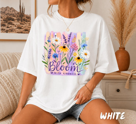 Inspirational floral graphic featuring colorful wildflowers including daisies, lavender, and black-eyed Susans over pastel watercolor brushstrokes with the quote "Bloom With Kindness" printed on a white unisex t-shirt.
