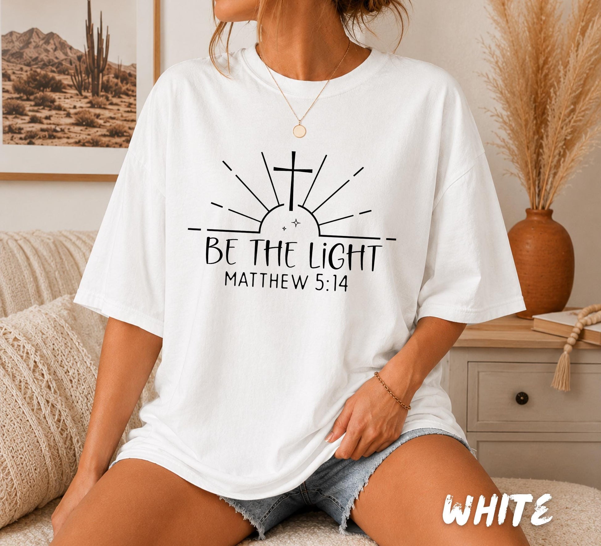 Woman wearing a white Comfort Colors Christian t-shirt featuring a minimalist "Be The Light" design with a cross, sunrise rays, and the Bible verse Matthew 5:14.
