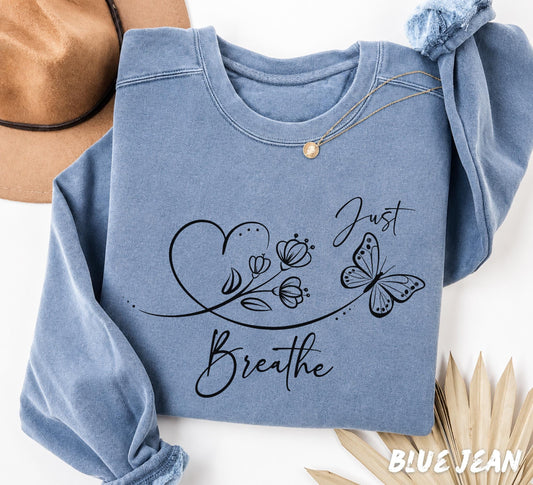Folded Blue Jean Comfort Colors sweatshirt displaying a minimalist "Just Breathe" design with delicate flowers, a butterfly, and flowing heart line art.