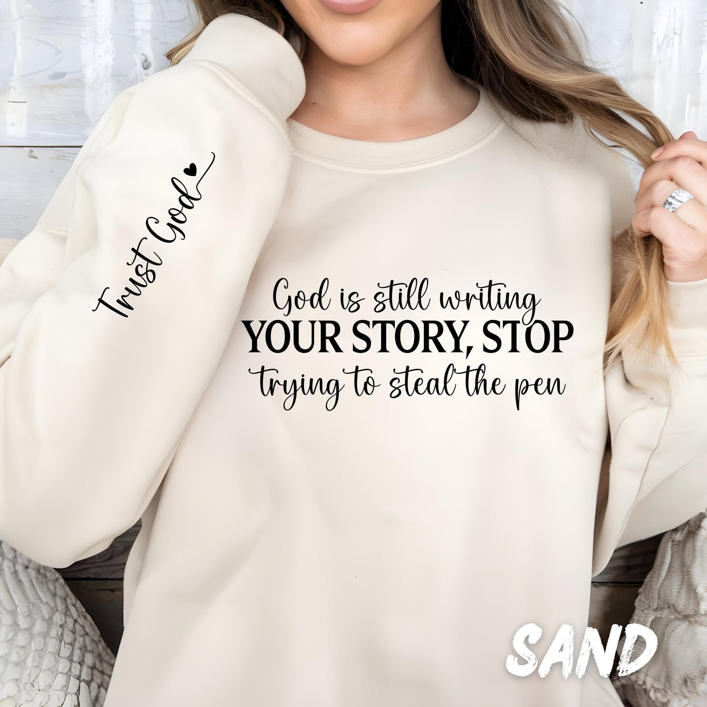 Sand Gildan crewneck sweatshirt featuring the inspirational "God Is Still Writing Your Story" design with a cross and colorful typography, plus a matching faith message printed on the sleeve.