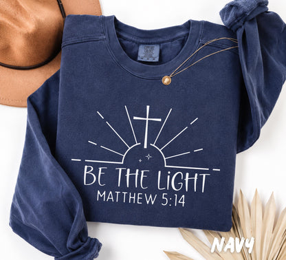 Folded Navy Comfort Colors crewneck sweatshirt featuring the inspirational Christian design "Be The Light" with Matthew 5:14, a cross, and minimalist sun rays.
