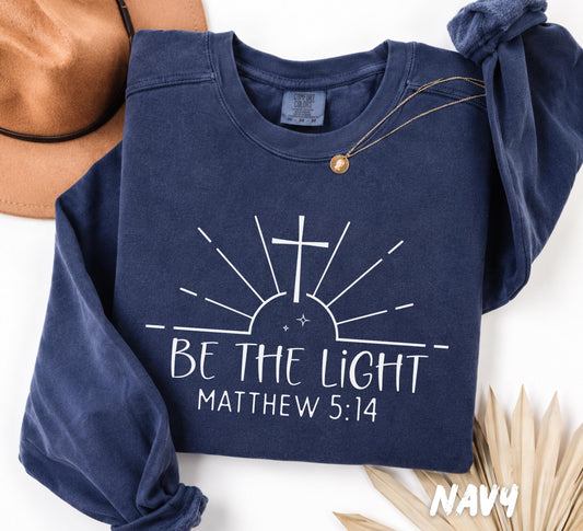 Folded Navy Comfort Colors crewneck sweatshirt featuring the inspirational Christian design "Be The Light" with Matthew 5:14, a cross, and minimalist sun rays.