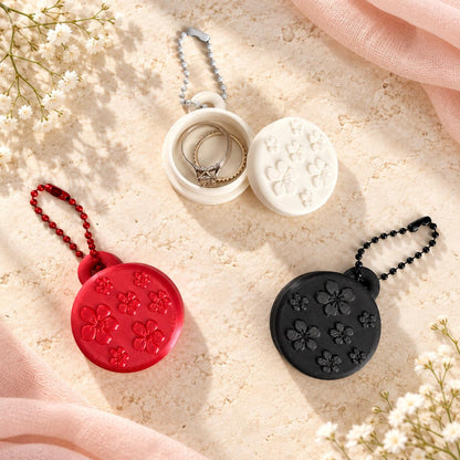 White, red, and black floral ring holder keychains displayed on a cream aesthetic background, with the white holder open to show the interior ring compartment.