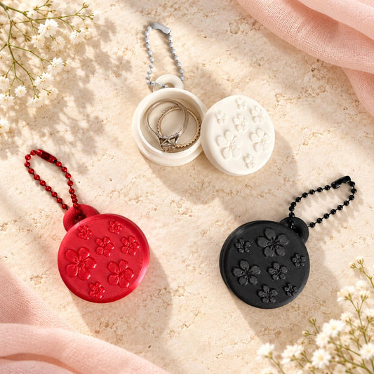 White, red, and black floral ring holder keychains displayed on a cream aesthetic background, with the white holder open to show the interior ring compartment.